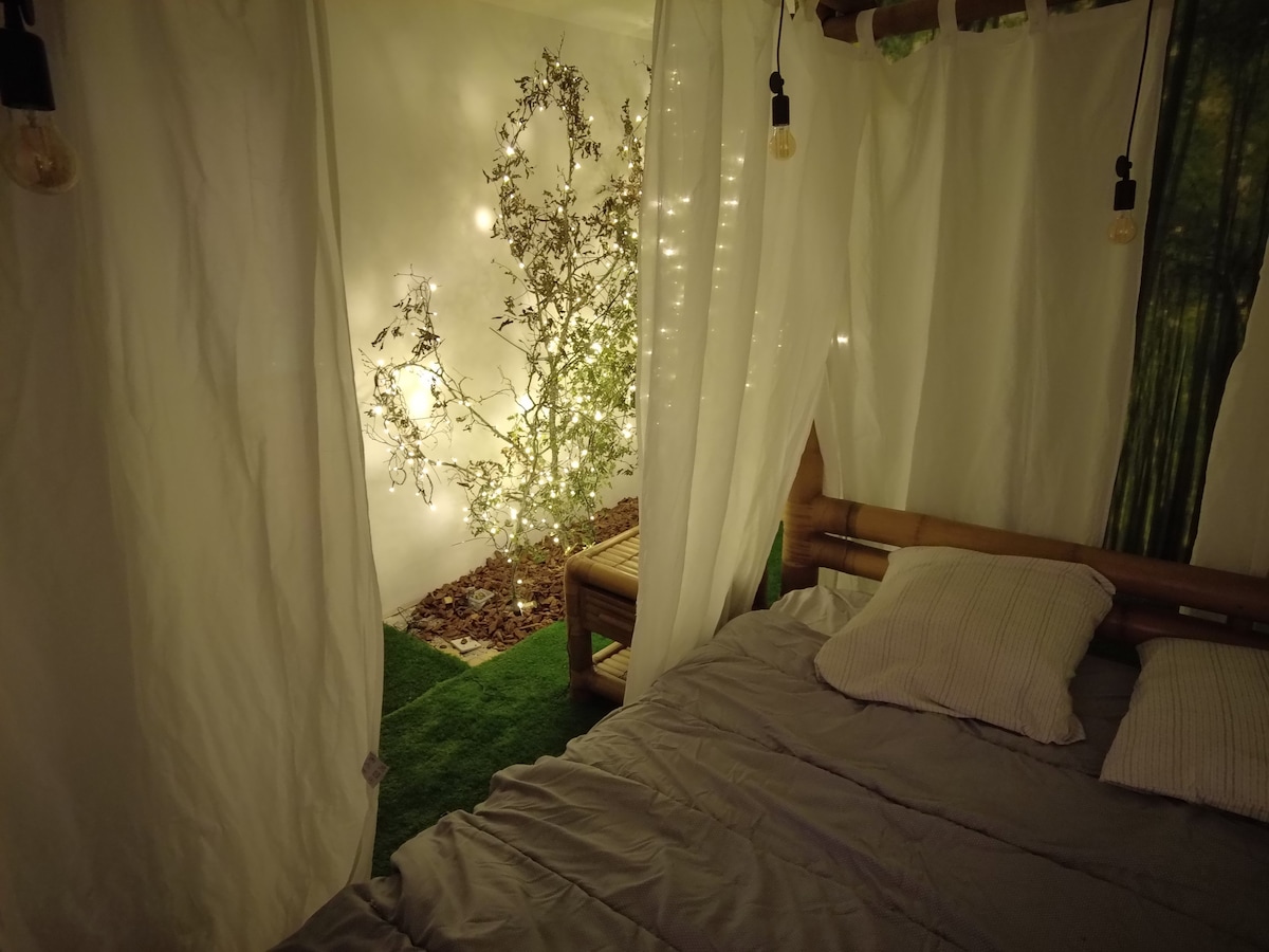 A serene sleeping area features a spacious bed with a light grey blanket and two pillows. Sheer white curtains create a cozy ambiance, framing an open space with a decorative tree adorned with soft lights, set against a green carpet of grass.