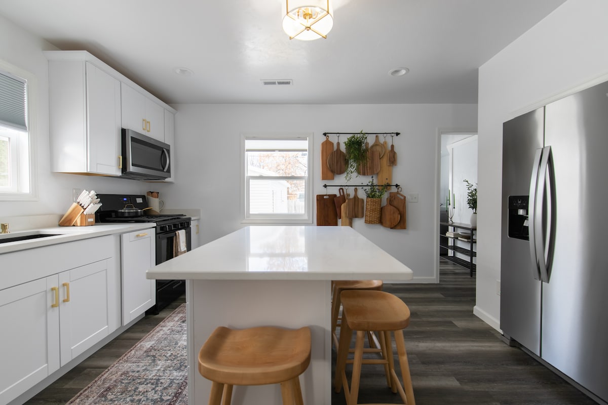 A modern kitchen with white cabinetry and stainless steel appliances features a large island with two wooden stools. Wall-mounted wooden boards add a touch of warmth. Natural light from the window highlights the sleek countertop and contemporary design.