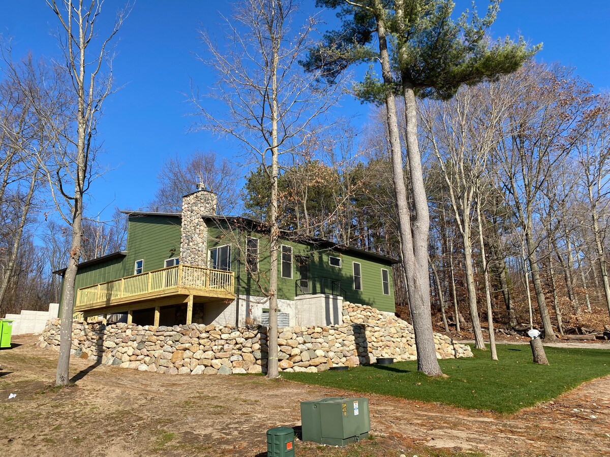 A newer green home is positioned on a stone foundation, featuring a wraparound deck and a chimney. Tall trees surround the property, with a clear blue sky above. Grass coverage is present, providing a natural setting for the inviting structure.