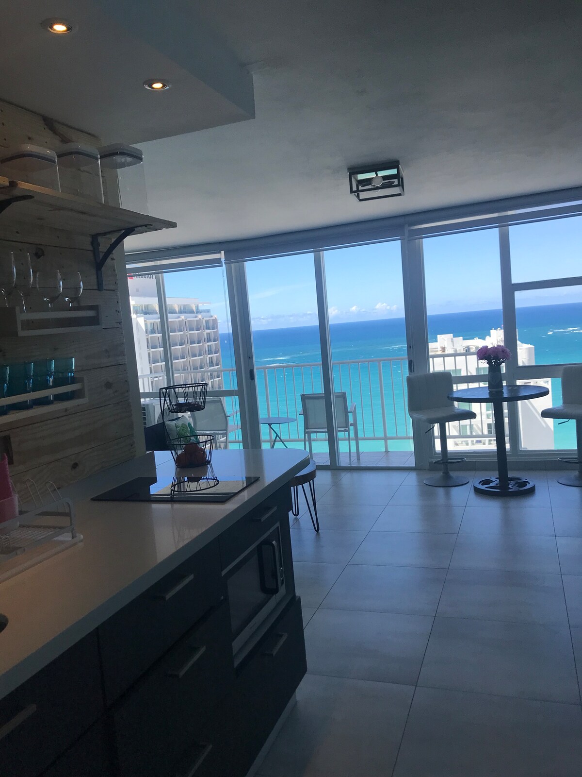The kitchen features a modern design with sleek cabinetry and a countertop. Large windows provide a panoramic view of the bright blue ocean, allowing natural light to fill the space. A dining area with chairs is visible, inviting moments of relaxation while enjoying the scenery.