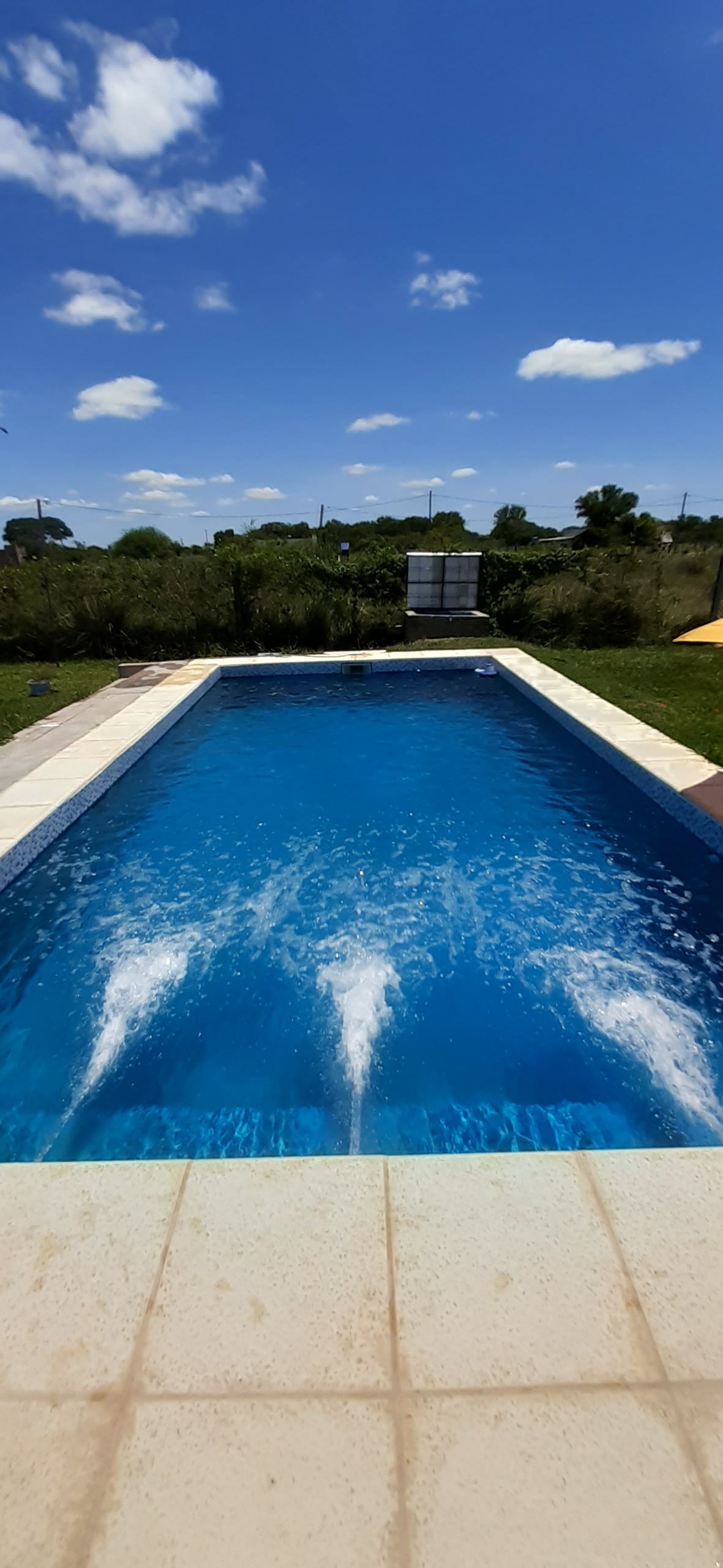 A clear blue pool is surrounded by a well-maintained lawn, with gentle water jets creating ripples across the surface. The bright blue sky and fluffy white clouds provide a serene backdrop to this outdoor space.