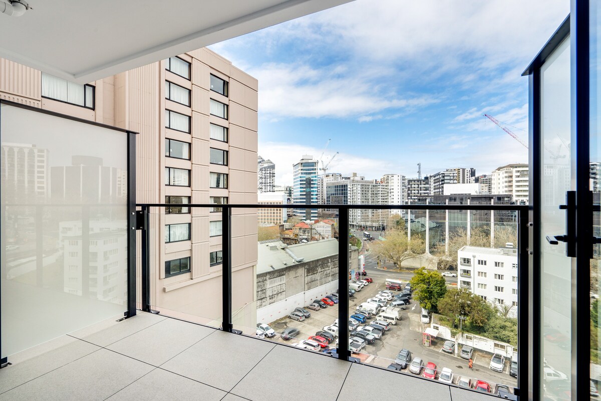 A balcony overlooks the vibrant cityscape of Auckland, with buildings featuring diverse architectural designs. The railing consists of clear glass panels, allowing for unobstructed views. Bright blue skies and scattered clouds create a welcoming outdoor atmosphere, inviting relaxation and enjoyment of the urban surroundings.
