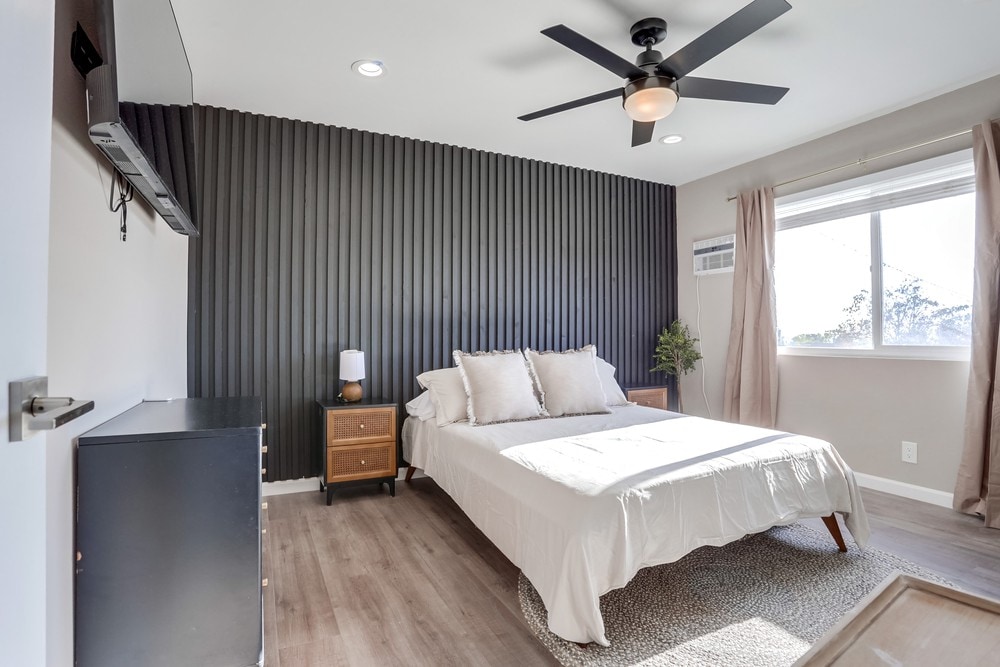 A spacious bedroom is highlighted by a light-colored bedspread over a wide bed. Soft lighting is provided by a bedside lamp. A modern ceiling fan is suspended above, while a window allows natural light to filter in, partially illuminating the textured accent wall.
