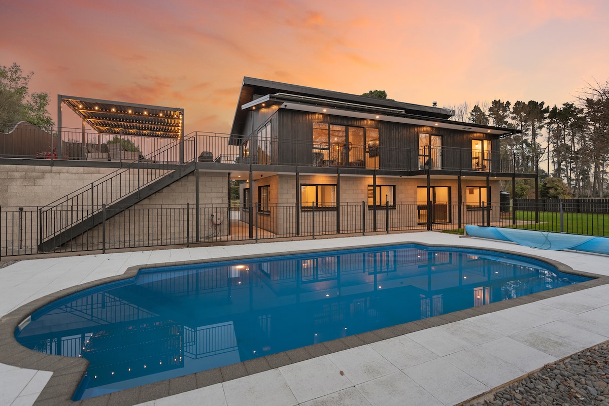 A modern house is set against a colorful sunset, with a spacious pool reflecting the sky. The two-story structure features large windows, allowing visibility of the interior lights. A patio area with string lights can be seen above, and expansive green space surrounds the property.