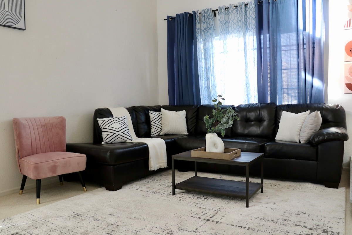 A spacious living area features a large black sectional sofa adorned with two decorative pillows. A light throw blanket is draped casually over the armrest. A chic pink chair complements the seating arrangement, while sheer blue curtains frame the window, allowing soft natural light to filter in.