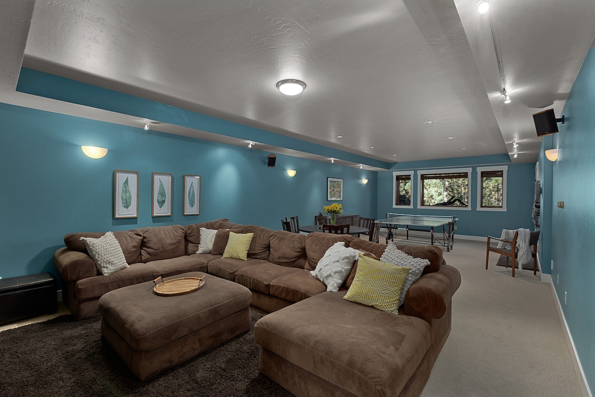 A spacious family room features a large wrap-around couch with decorative pillows. A ping pong table is visible in the background, alongside a dining table and vibrant artwork on the walls. Natural light filters in through large windows overlooking the greenery.