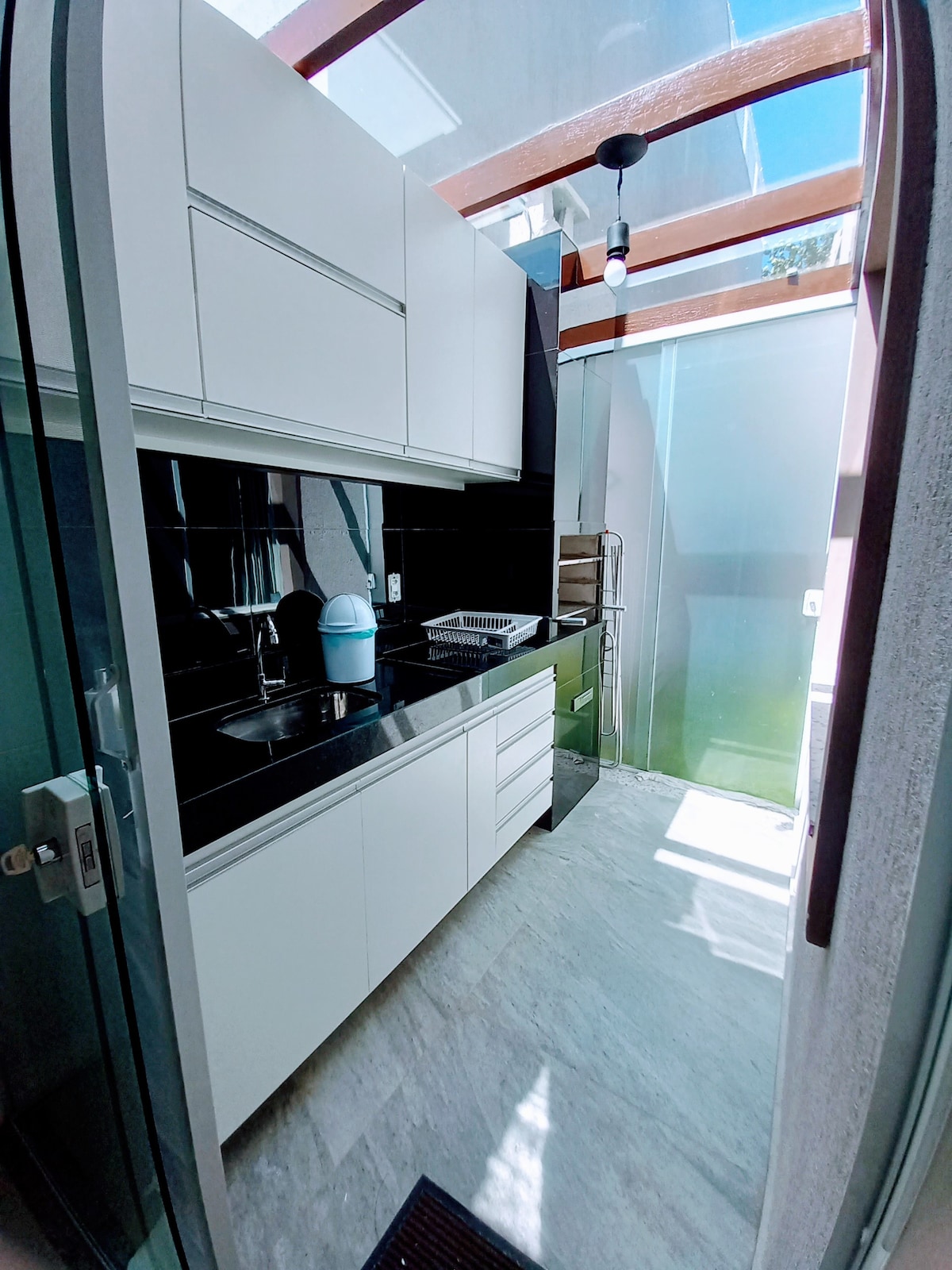 A modern kitchen space is featured, equipped with sleek white cabinets and a black countertop. A sink and stovetop are included, with glass walls allowing natural light to illuminate the area. The flooring is made of light-colored tiles, contributing to a clean and functional design.