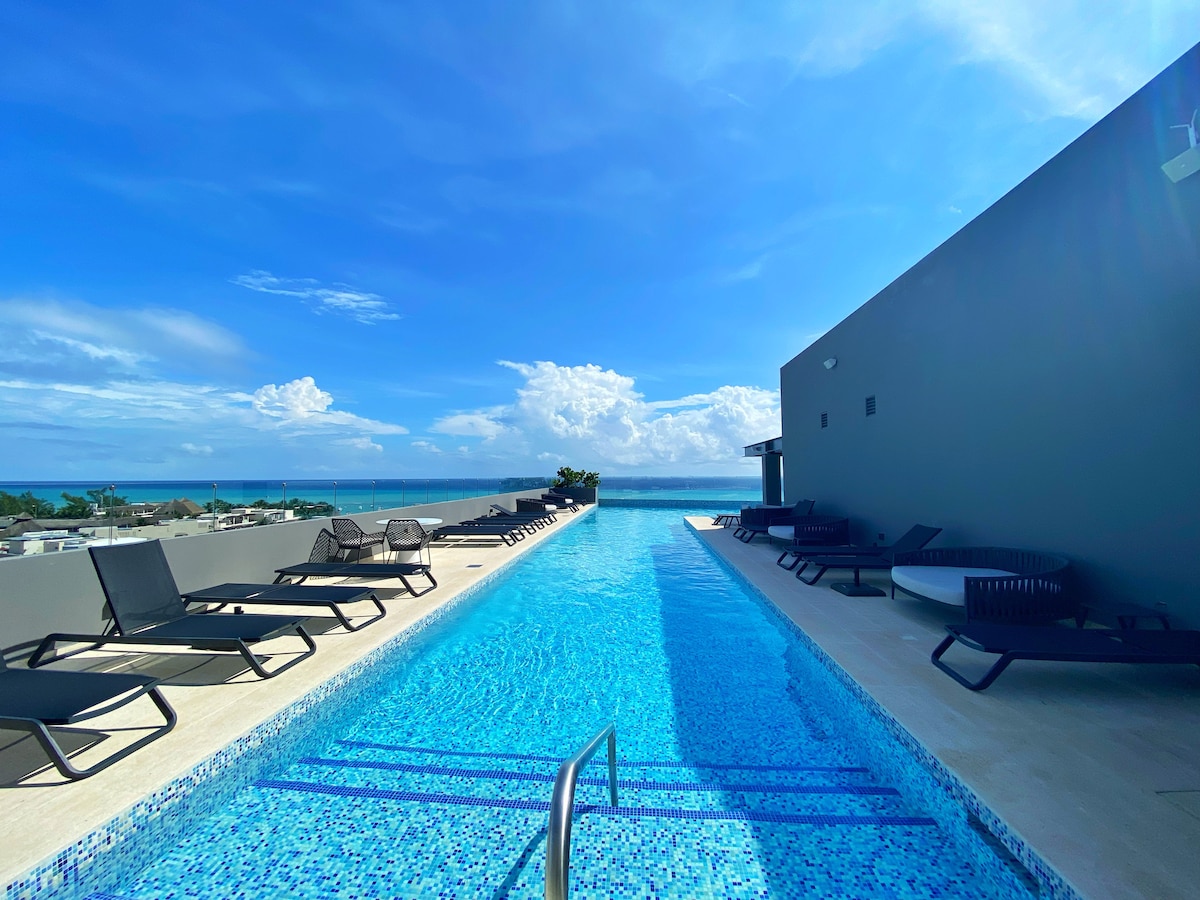 The rooftop pool is depicted with vibrant blue water reflecting the sky. Comfortable lounge chairs are arranged along the pool's edge, providing scenic views of the ocean and surrounding landscape. The serene atmosphere is complemented by the clear blue sky, accentuating the tranquil setting.