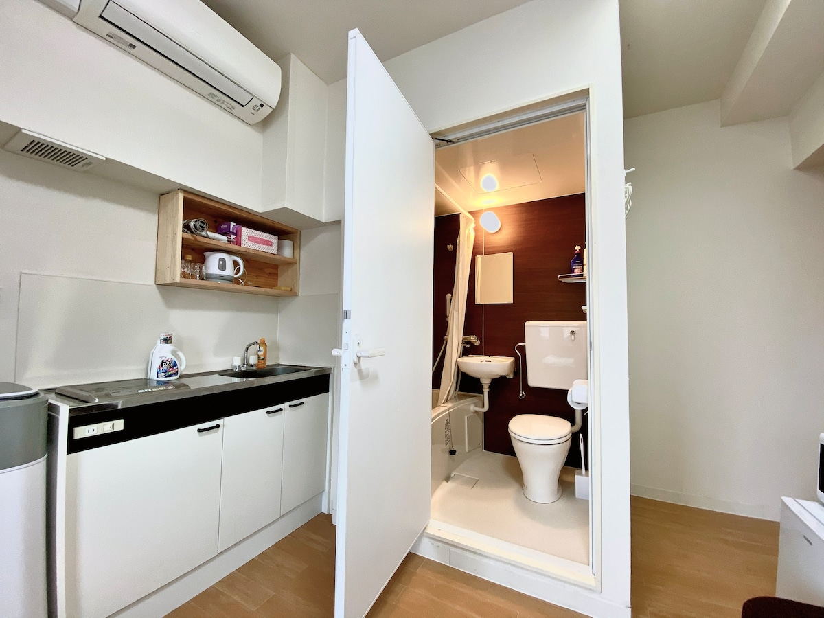 A functional kitchenette is visible with essential appliances, including a microwave and refrigerator. A partially open door reveals an ensuite bathroom featuring a toilet and shower area. The overall space is well-organized, with light walls enhancing the ambiance.