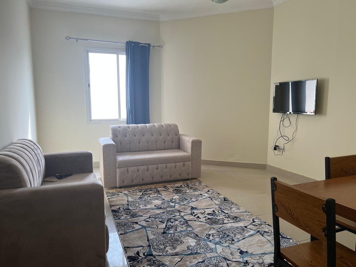 A living area is presented with two seating arrangements, featuring a beige couch and a light-colored loveseat. A flat-screen television is mounted on the wall opposite a small dining table with chairs. Natural light enters through a window draped with dark curtains.