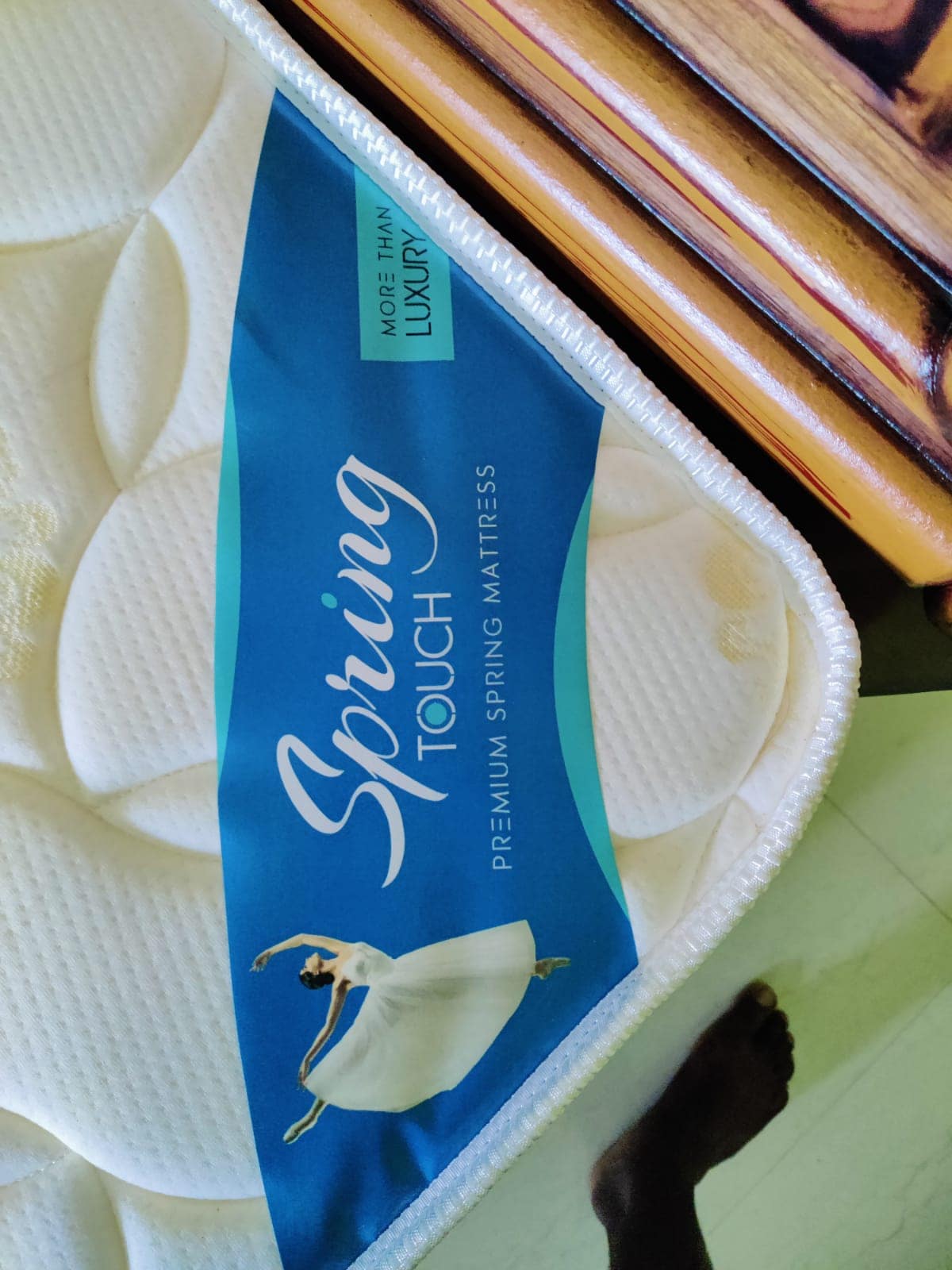 A close-up of a premium spring mattress is displayed, featuring a blue label with elegant lettering. The mattress surface is lightly textured, and a corner of a wooden bedside table and part of a foot are visible in the frame.