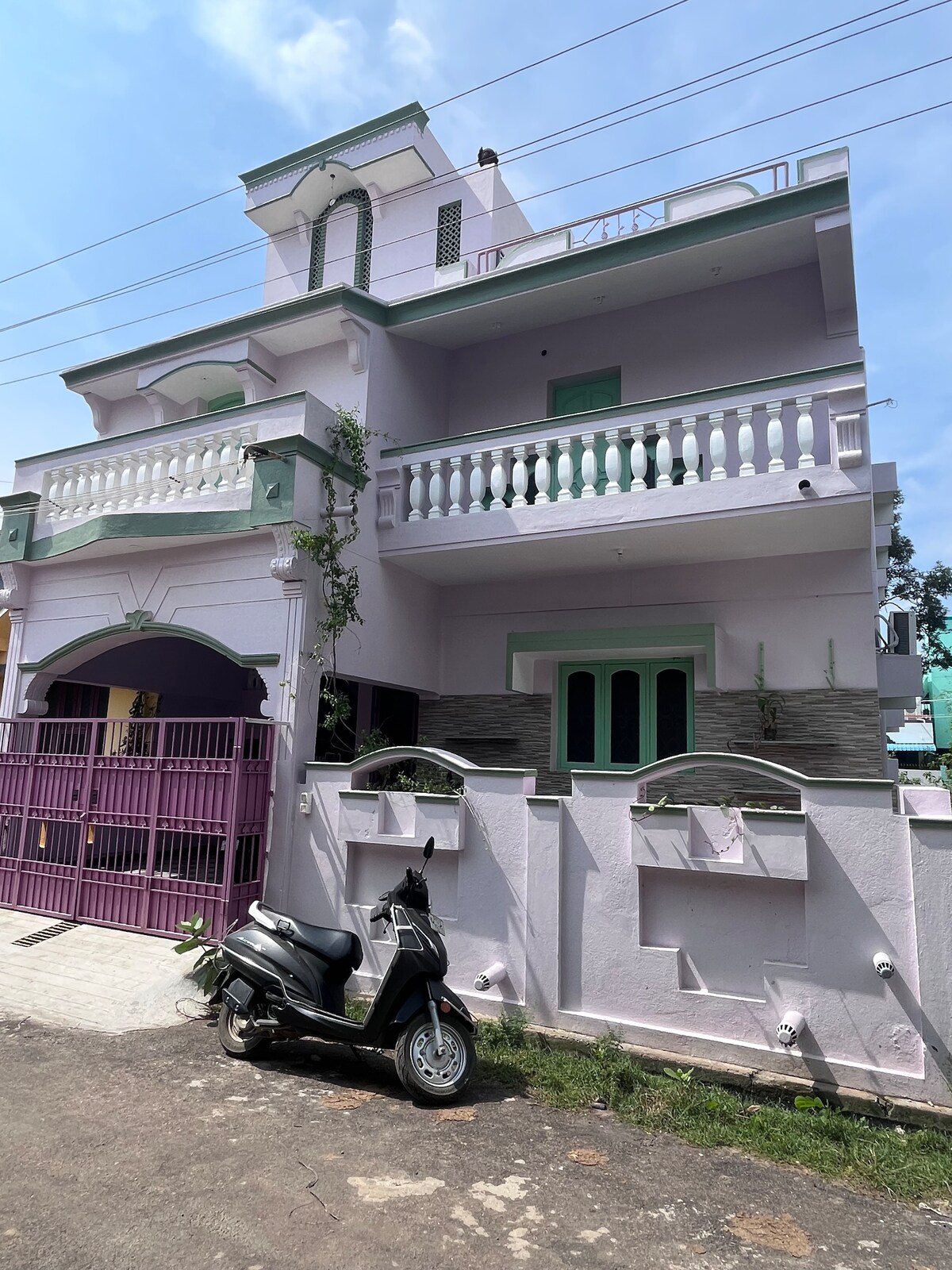 A multi-story house is depicted with a light purple façade, featuring a balcony adorned with white railings. The entrance is framed by a purple gate. A parked scooter is visible in the foreground, with manicured greenery complementing the property.