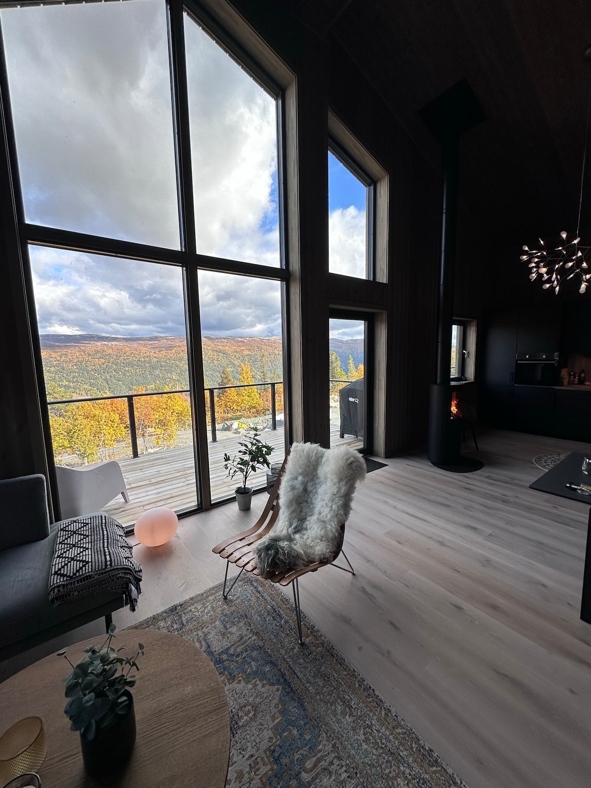 A spacious living area features large floor-to-ceiling windows offering expansive views of the surrounding landscape. A cozy chair with a soft throw is placed near a small table with a plant. A fireplace is visible in the background, adding warmth to the atmosphere.