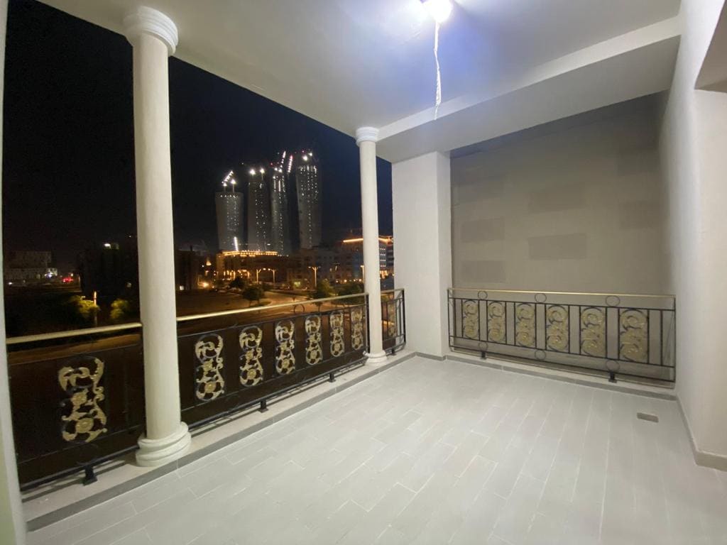 A spacious balcony features elegant railings, framed by tall columns. The city skyline is visible in the background against a night sky, with illuminated buildings adding a dynamic touch to the scene. The smooth tiled floor enhances the openness of the space.