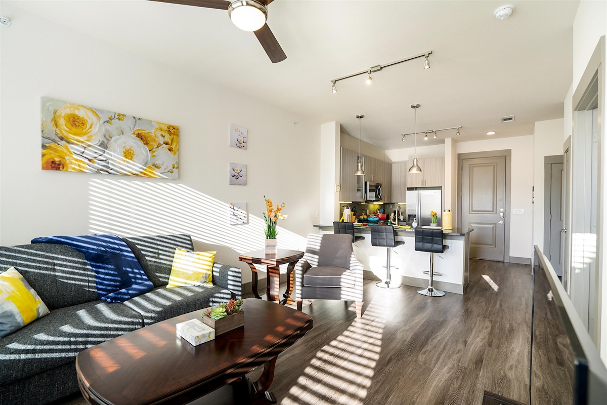 A modern living space features a cozy sofa adorned with bright pillows and a wooden coffee table. Sunlight streams through the window, creating a pleasant ambiance. The open kitchen showcases sleek countertops and bar seating, complemented by contemporary decor and a floral wall art.