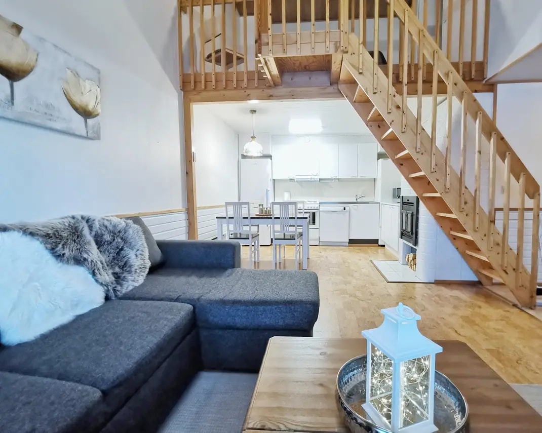 A spacious living area features a comfortable dark grey sofa with a fluffy throw, positioned beside a wooden coffee table. A staircase leads to the upper level, while a dining area with a table and chairs is visible in the background, adjacent to a modern kitchen.