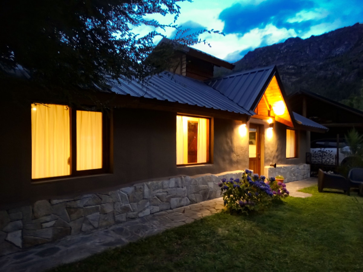 The exterior of the cabin is shown at twilight, featuring a stone foundation and wooden accents. Soft lighting illuminates the entrance and window areas. The well-maintained garden includes flowering plants, with a backdrop of mountains visible in the evening sky.