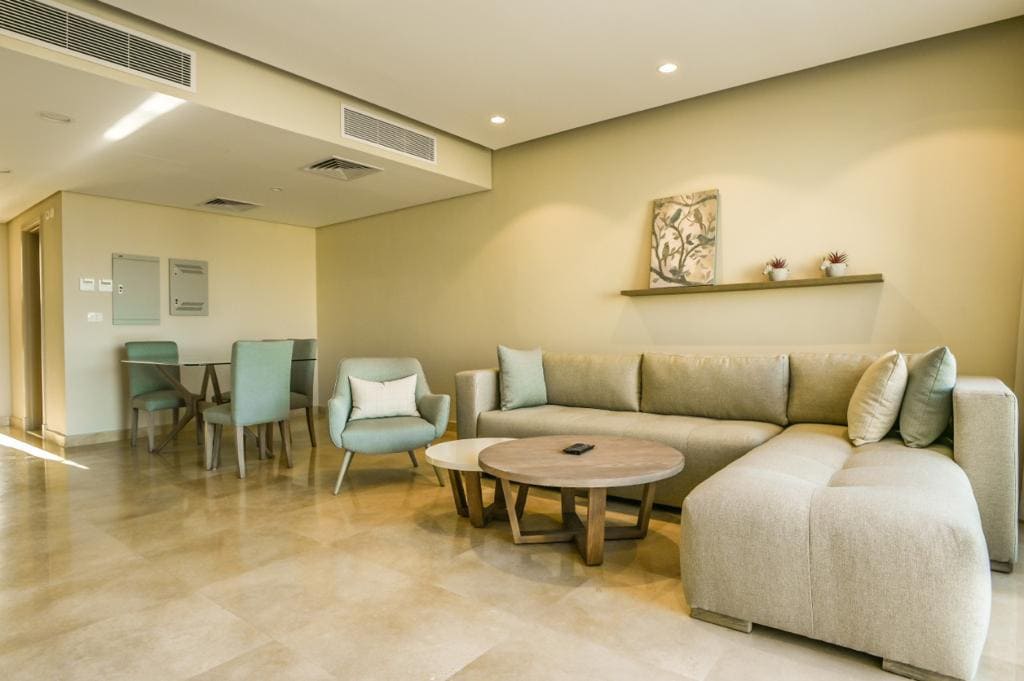 A spacious living area is furnished with a light-colored sectional sofa and a round wooden coffee table. Several chairs with soft upholstery complement the sofa. A wall shelf displays a decorative piece, while warm lighting enhances the neutral tones of the room.