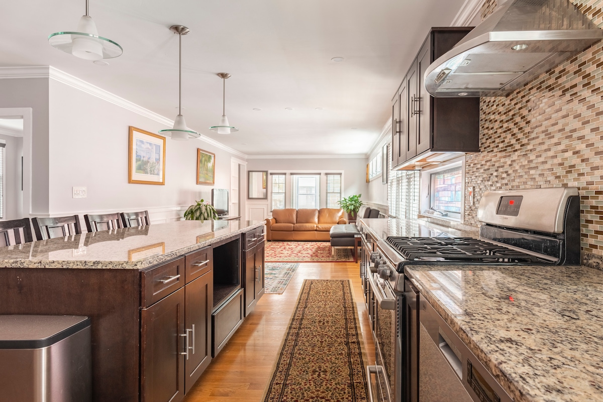 A fully equipped kitchen features modern stainless steel appliances and granite countertops. An island with seating is positioned along one side, with a cozy living area visible in the background. Natural light fills the space, highlighting the warm wood flooring and contemporary design.