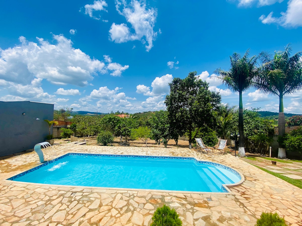 An inviting outdoor space features a swimming pool surrounded by stone paving. Lush greenery and palm trees are visible in the background, providing a serene atmosphere. Clear blue skies create a bright and cheerful setting for relaxation.