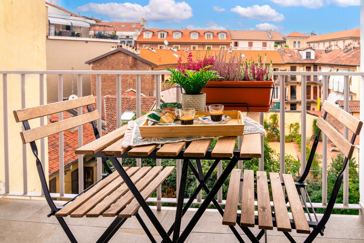 A small balcony features a wooden table and two chairs, set with a tray of beverages. Potted plants are displayed, providing a touch of greenery. The view shows rooftops of nearby buildings under a clear blue sky.