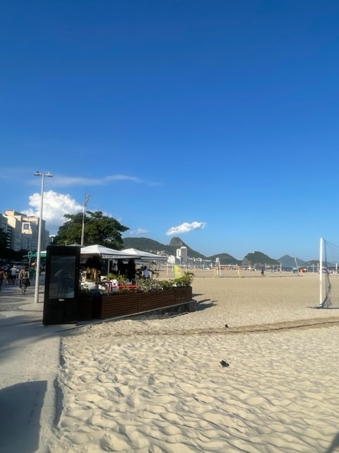 A beachfront view showcases a sandy area leading to the ocean, with a vendor stand featuring a white canopy and greenery. Mountains rise in the distance under a clear, blue sky, highlighting the vibrant atmosphere of the beach setting.