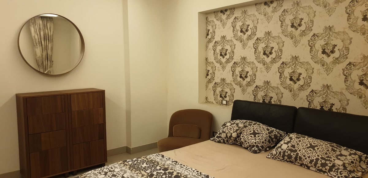A modern bedroom features an upholstered headboard with decorative bedding. A round mirror hangs above a wooden dresser, and a cozy chair is positioned beside the bed. The wallpaper showcases an intricate pattern, contributing to the room's stylish ambiance.