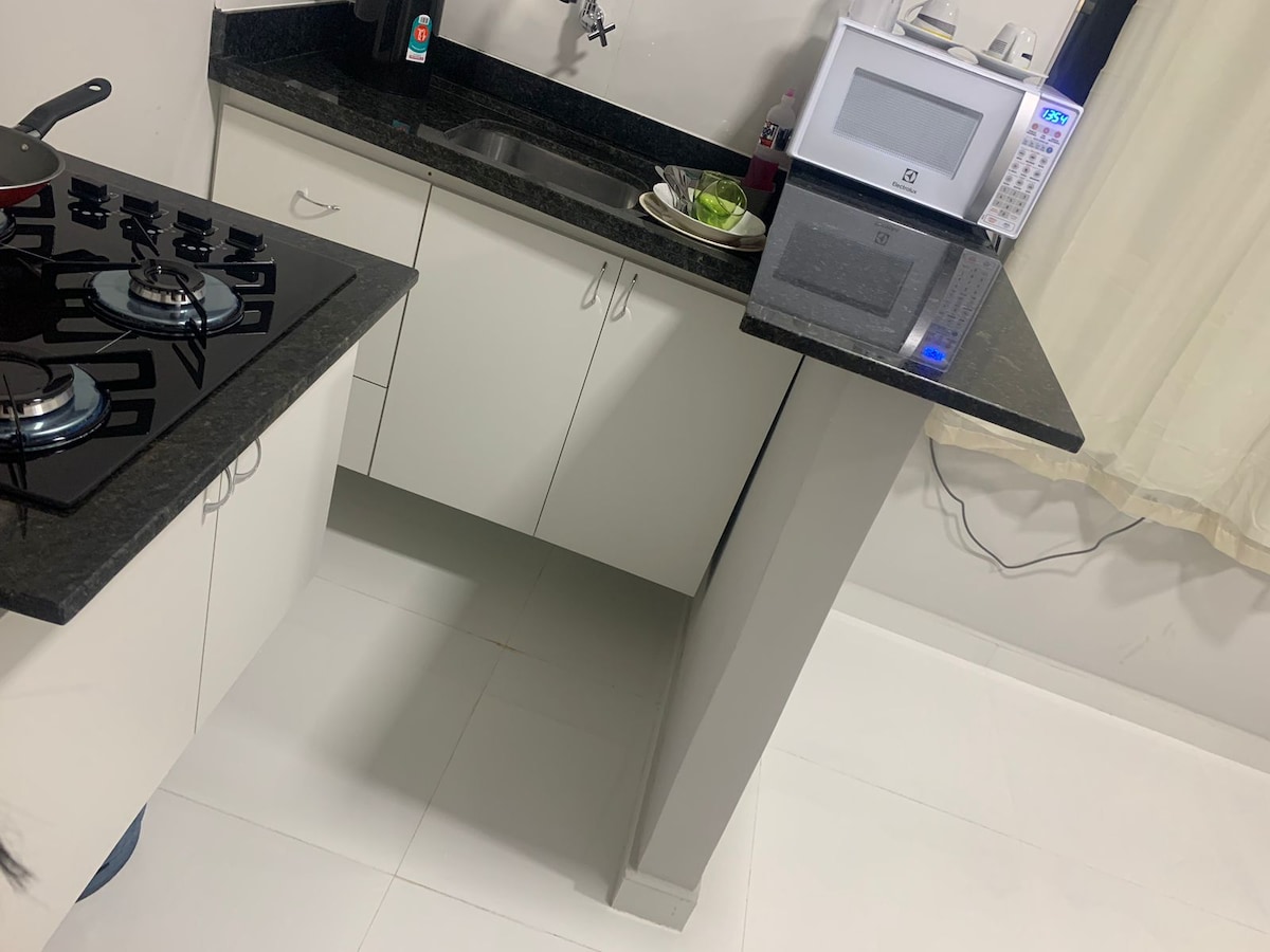 A modern kitchen area is shown featuring a black granite countertop. A four-burner cooktop is visible along with a microwave and various kitchen utensils. A sink is positioned near the counter, while natural light enters through sheer curtains.