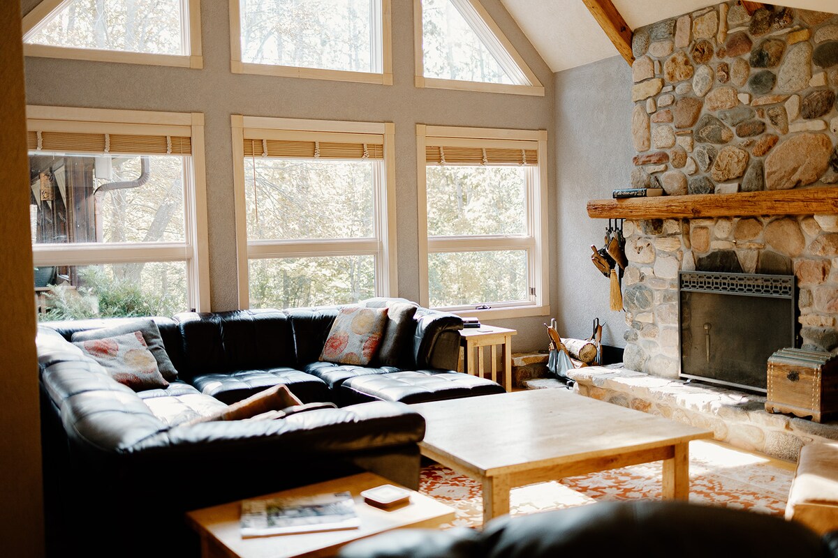 A spacious living area is featured with a large, dark sectional sofa arranged around a wooden coffee table. Sunlight filters through expansive windows, illuminating the room. A stone fireplace adds warmth and character, while soft area rugs contribute to a cozy atmosphere.