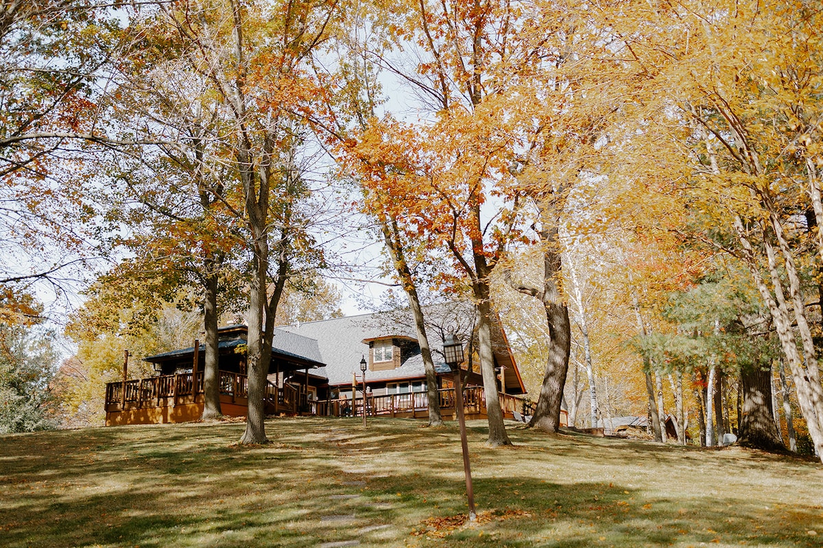 The ranch-style retreat is surrounded by autumn foliage, with vibrant orange and yellow leaves visible on the trees. The structure is enhanced by a spacious wrap-around porch, providing an inviting outdoor area that blends harmoniously with the natural surroundings.