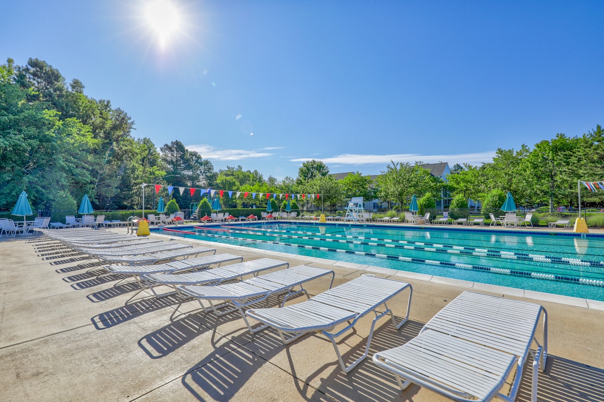 An outdoor pool area is depicted, featuring a long swimming pool lined with lounge chairs. Bright sunlight casts a warm glow over the scene, while colorful flags are strung across the pool. Lush greenery surrounds the area, adding to the inviting atmosphere.