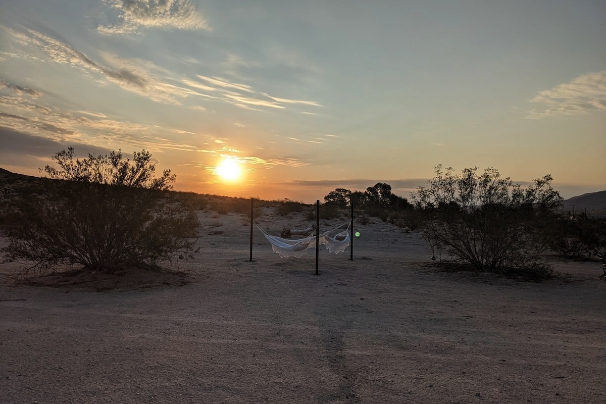A serene desert landscape features a pair of hammocks suspended between sturdy poles, positioned to catch the warm glow of the setting sun. Surrounding shrubs and the open terrain create a peaceful atmosphere as the sun dips below the horizon.