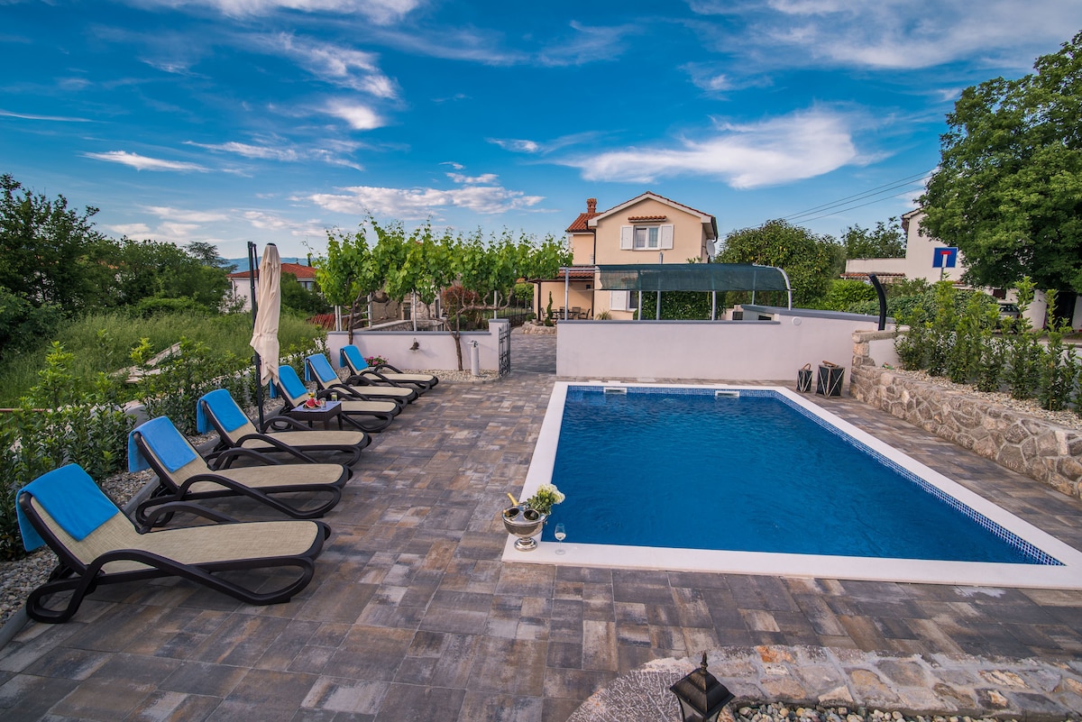 A private pool is surrounded by a spacious sunbathing area featuring several lounge chairs. The area is lined with greenery and a view of a two-story villa is visible in the background, under a blue sky with scattered clouds.