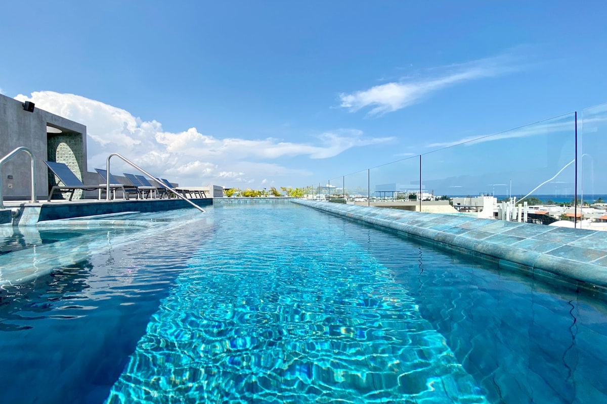 An expansive rooftop pool glistens under the sun, surrounded by clear glass railings. The water reflects a bright blue sky, offering views of the ocean and skyline. Comfortable lounge chairs are positioned nearby for relaxation.