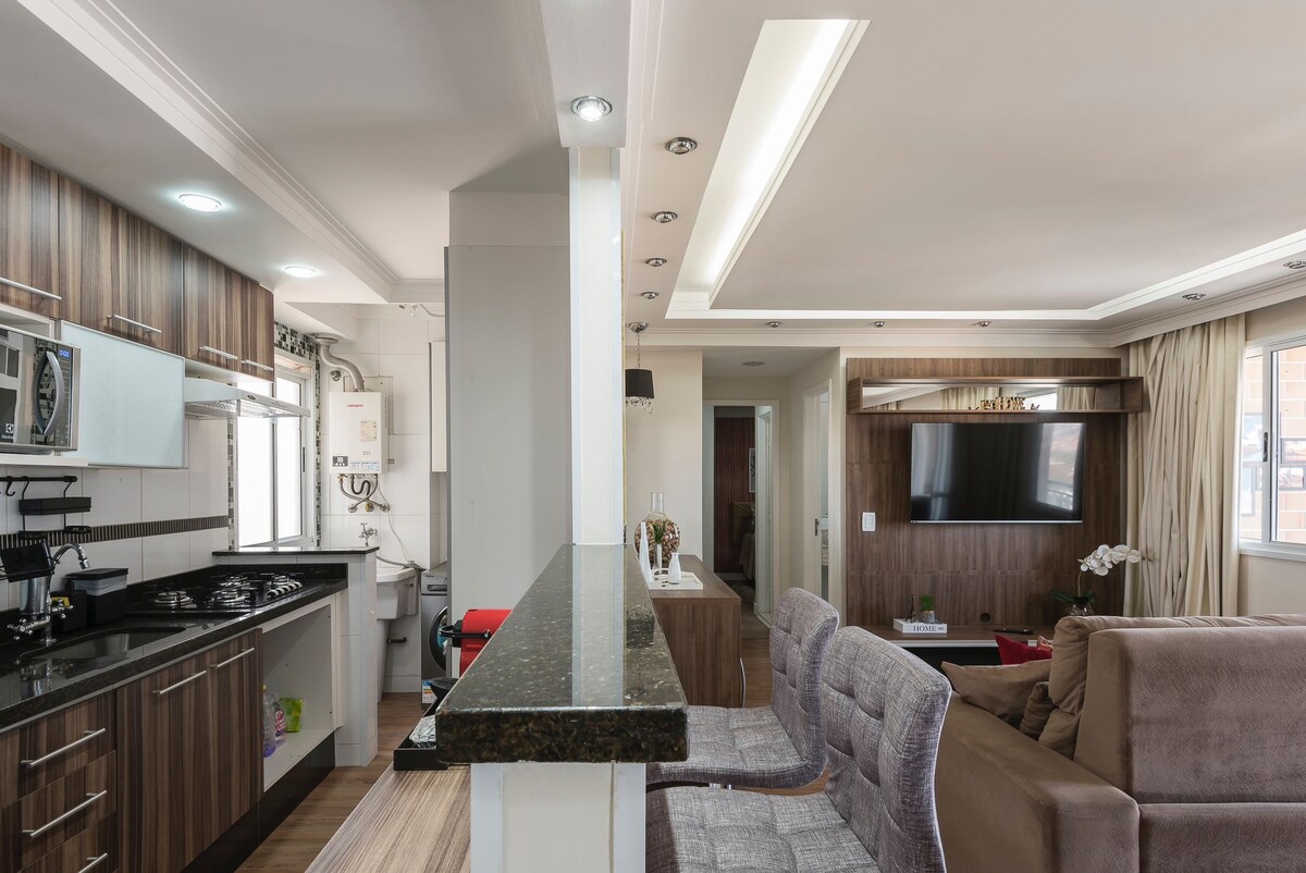 The open-plan living area features a modern kitchen with dark cabinetry and a countertop that overlooks seating arrangements. Light floods in through large windows, highlighting the comfortable sofa and entertainment center with a flat-screen TV, creating a welcoming space for relaxation.