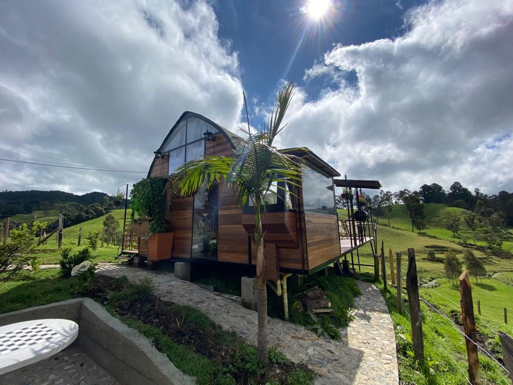 A charming wooden cabin is set amidst rolling green hills, featuring large windows that offer views of the landscape. A small palm tree is visible in front, while a deck extends from the structure, inviting outdoor relaxation. The sky is partly cloudy with a bright sun overhead.