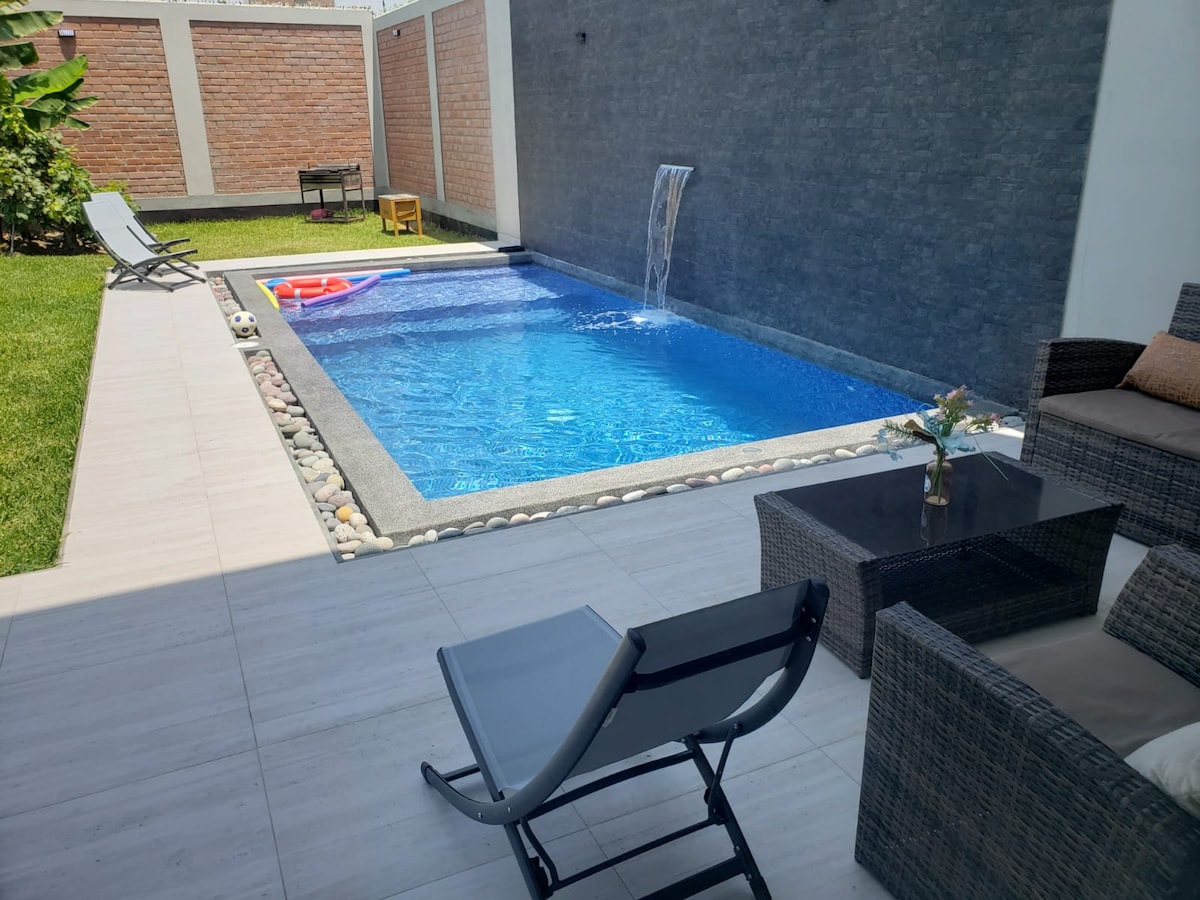 A tranquil outdoor area features a swimming pool with a water feature, surrounded by a stone border. Lounge chairs are positioned beside the pool. A small seating arrangement is present on the patio, accompanied by a landscaped area featuring grass and greenery.