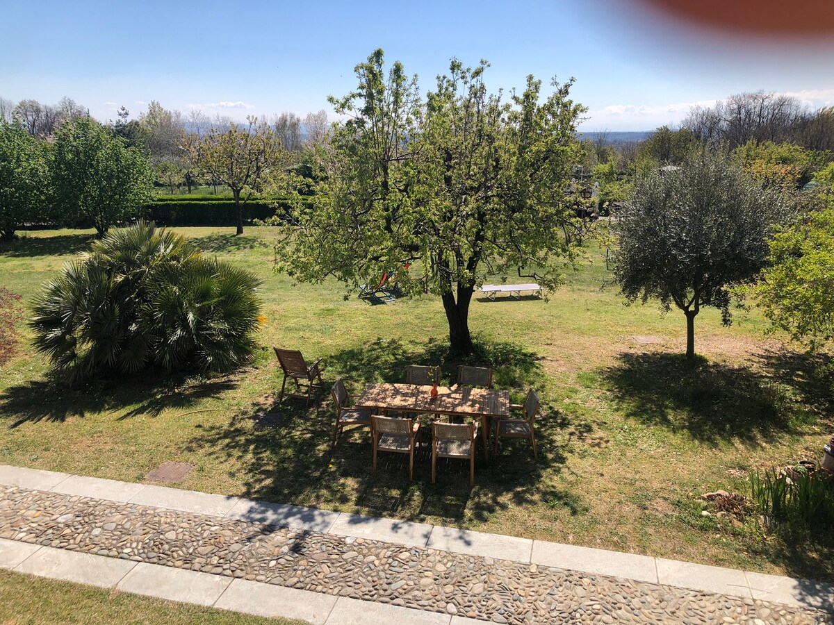 An outdoor area is presented, featuring a wooden table surrounded by several chairs. Lush greenery, including trees and plants, borders the space, enhancing the natural setting. A well-maintained lawn stretches toward the horizon, providing a peaceful environment for outdoor activities.