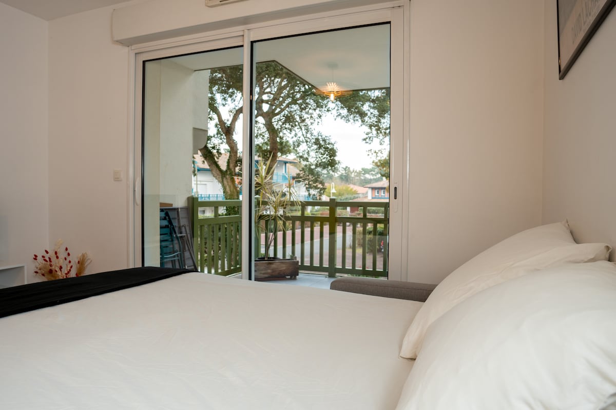 A spacious bed is positioned in front of large sliding glass doors that open to a balcony. The outdoor area is surrounded by greenery, providing a serene view. Soft natural light enhances the inviting atmosphere of the room.