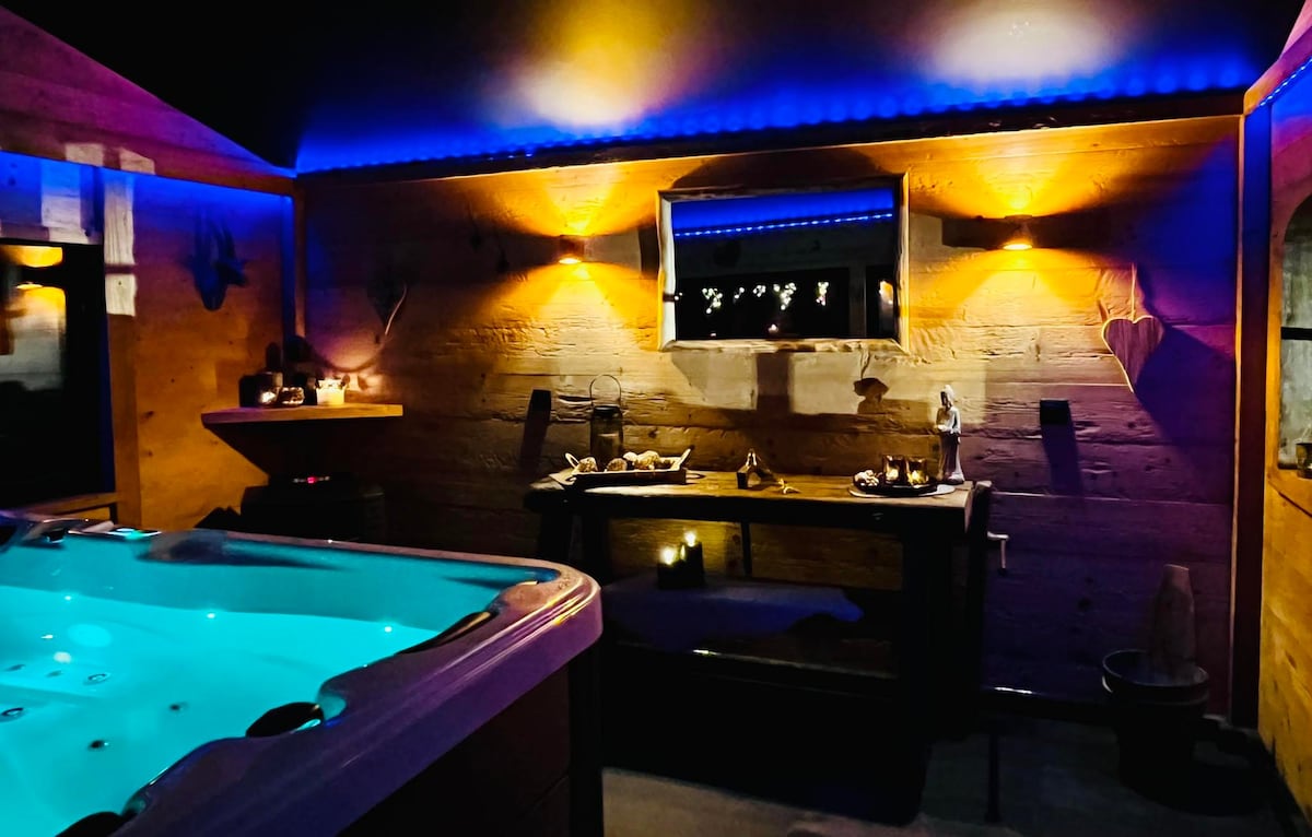 An elegantly designed spa area features a jacuzzi with soft blue lighting, creating a serene ambiance. Wooden walls are adorned with subtle decor, and calming candlelight illuminates the space. A counter displays various amenities, enhancing the tranquil environment for relaxation.