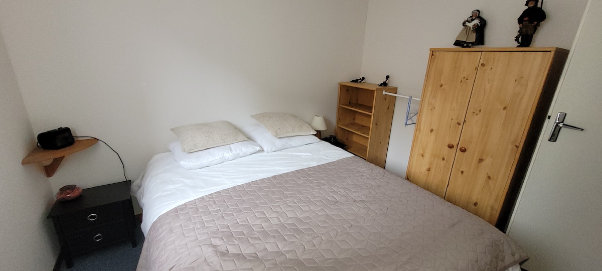 A well-lit sleeping area features a spacious bed with soft white linens and beige accent pillows. A wooden wardrobe and bookshelves provide storage, while a small bedside table holds a lamp. Decorative figurines are displayed on top of the wardrobe.