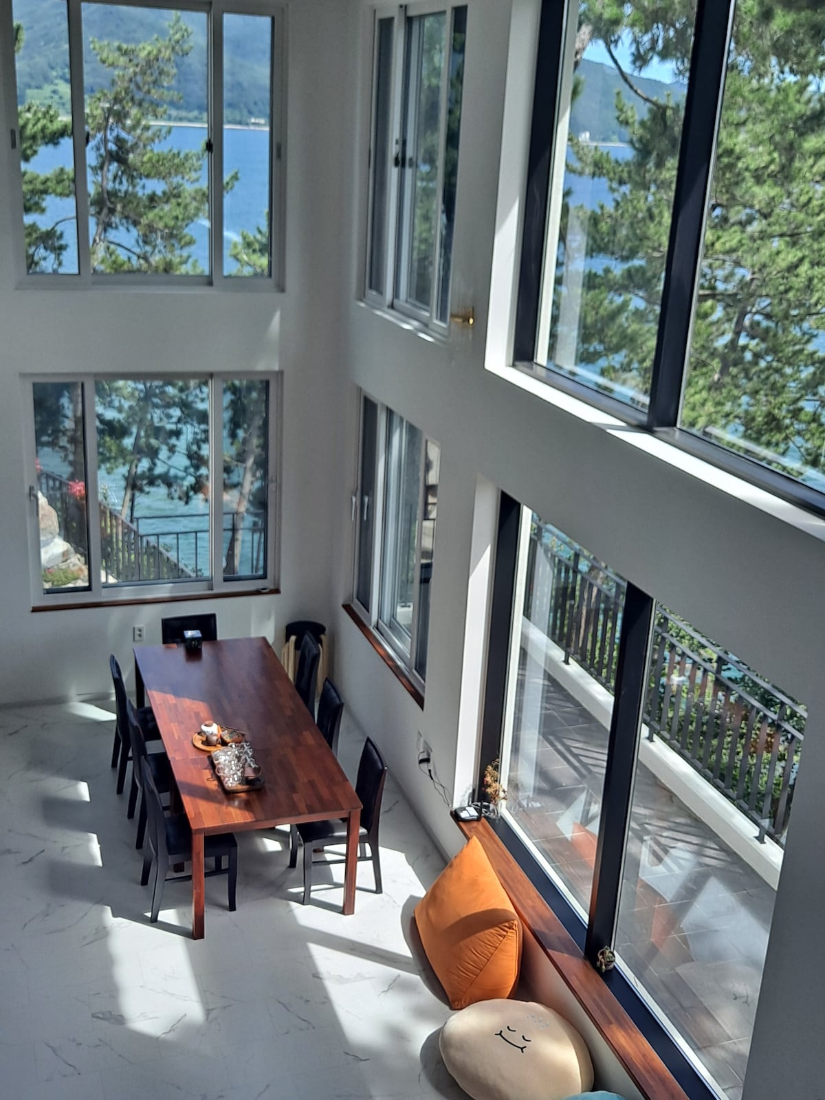 A spacious dining area features a long wooden table surrounded by black chairs. Large windows provide abundant natural light and a view of the trees and water outside. A cozy seating area includes a large cushion by the window.