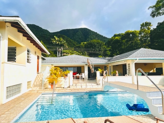 A private pool is set within a spacious outdoor area, surrounded by lush greenery and mountains in the background. Comfortable lounge chairs line the poolside, with a patio dining area visible behind. Sunlight filters through the trees, creating a serene atmosphere.