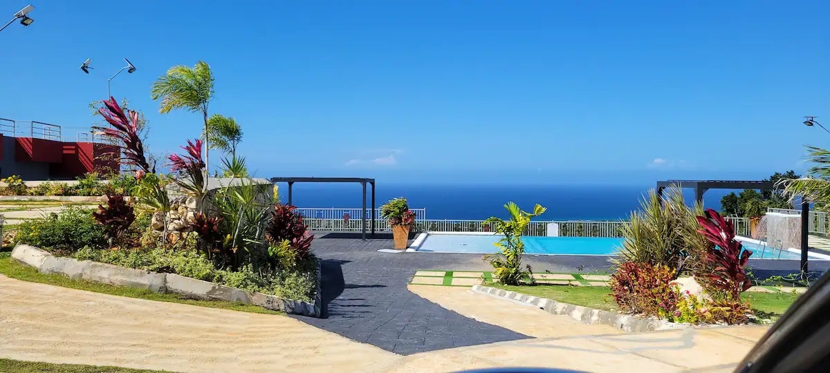Lush landscaping surrounds a serene pool area, with vibrant tropical plants and shrubs lining the pathway. A clear blue ocean stretches into the distance under a bright sky, enhancing the inviting atmosphere of the outdoor space.