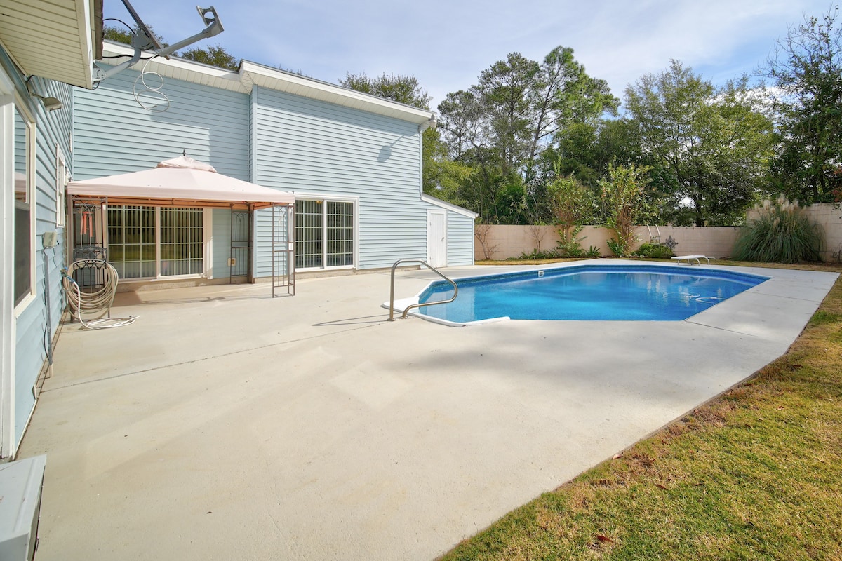 The private backyard features a shimmering pool bordered by a spacious concrete patio. A shaded seating area is provided by a light-colored gazebo. Lush greenery and a tall wall ensure privacy, creating a serene outdoor environment.