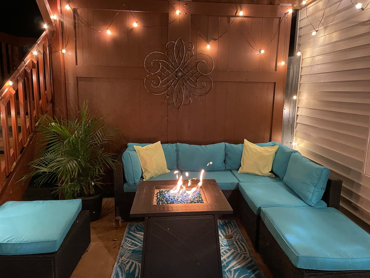 A comfortable outdoor seating area features sectional seating with blue cushions arranged around a fire table. Soft lighting from string lights enhances the ambiance. A potted plant adds a touch of greenery, creating a cozy corner for relaxation.