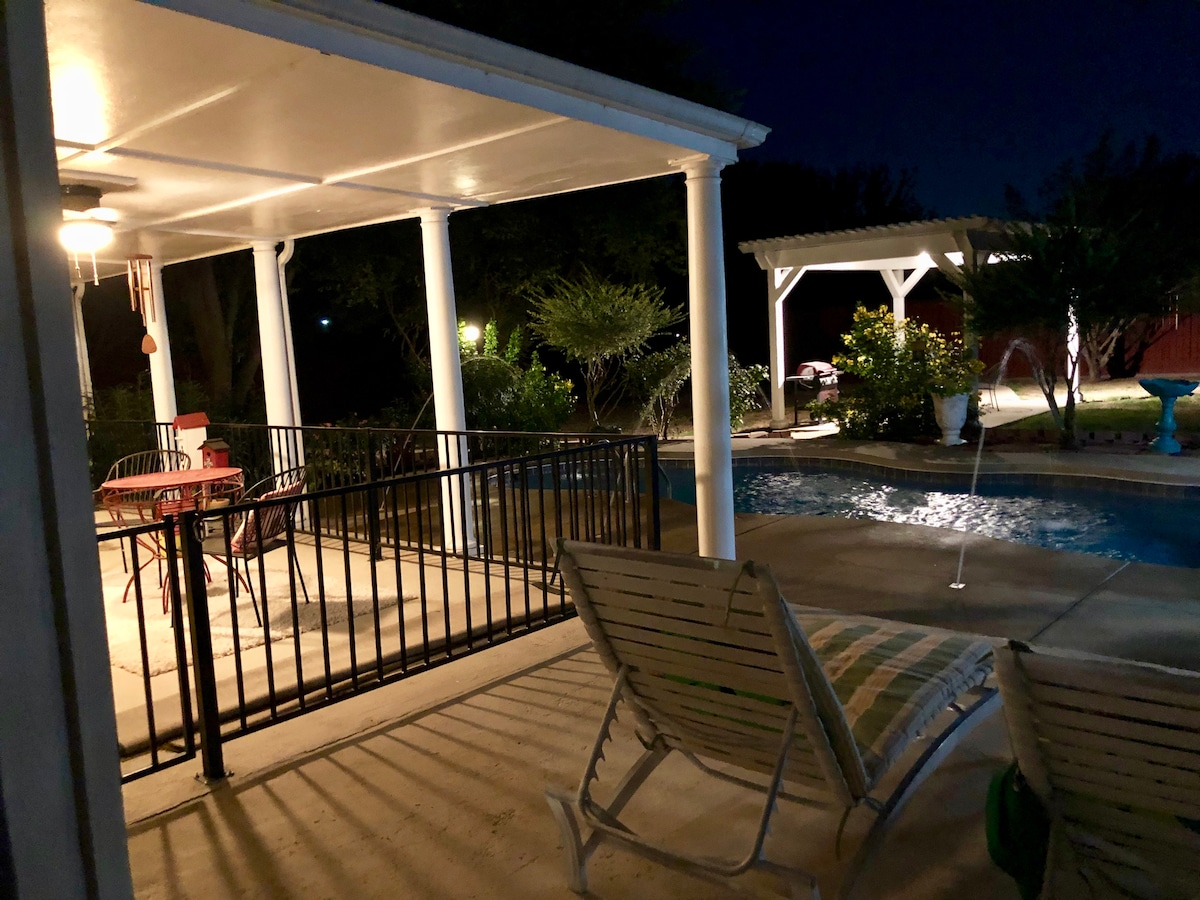 A well-lit outdoor area features seating arrangements near a pool, with lounge chairs positioned beside the water. A covered patio provides shade, while nearby greenery adds a natural touch. Subtle lighting enhances the space, creating a relaxing atmosphere for evening gatherings.
