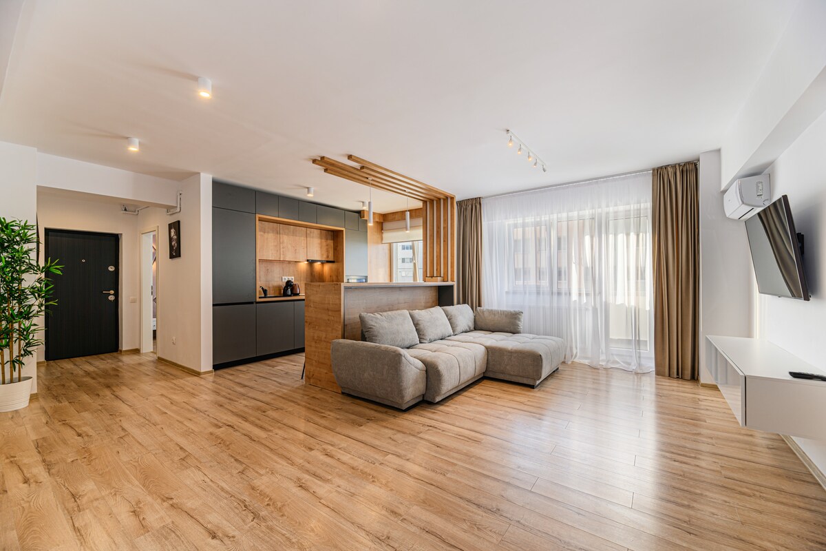 A spacious living area features a modern L-shaped sofa and a sleek kitchenette with wooden accents. Natural light fills the room through the large window, which is dressed with light curtains. The wooden flooring complements the contemporary design.
