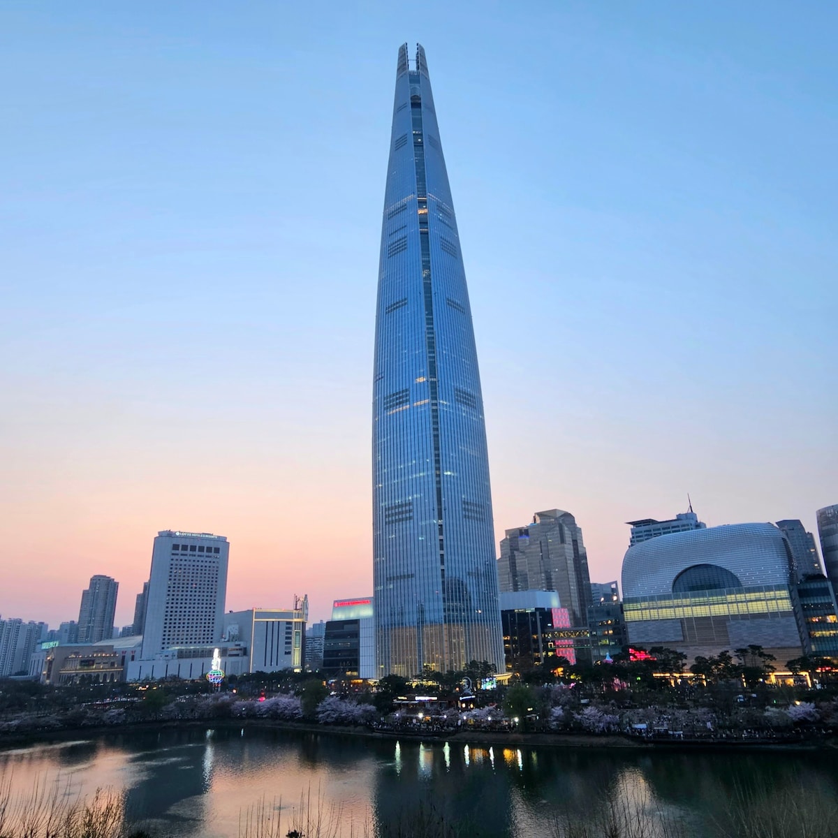 The Lotte World Tower stands tall against a pastel sky, reflecting shades of blue and pink. Surrounding buildings are visible, with the tranquil waters of a nearby lake mirroring the tower's impressive silhouette. Soft lights illuminate the base, enhancing the city's evening ambiance.