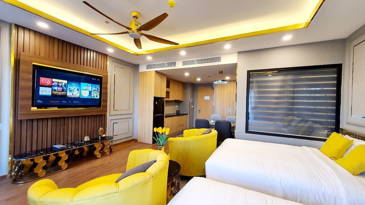 A modern living space features two spacious beds complemented by bright yellow accents. A large flat-screen TV is mounted on the wall, with a cozy seating area consisting of two circular chairs. Natural light filters through the window, enhancing the warm wood tones.